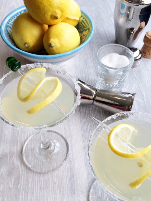 A set up for making lemon cocktails. A bowl of lemons, shot class of sugar, 2 martini glasses full of cocktail with a lemon twist on a skewer.
