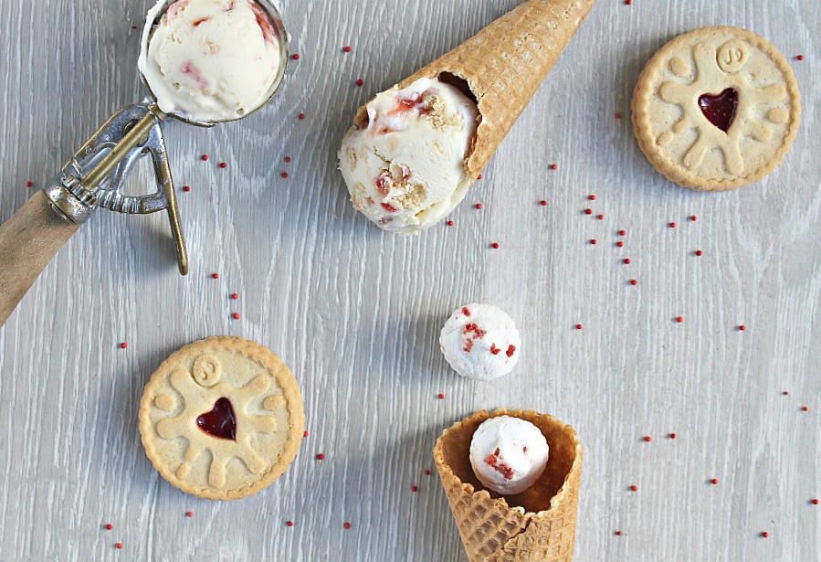 An ice cream scoop with one scoop of ice cream in it, 2 waffle cones, 1 has ice cream and one has a marshmallow, and 2 biscuits all on a table