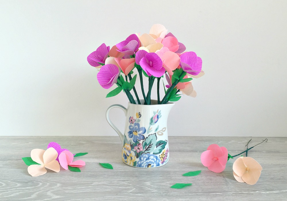 A jug with a bouquet of crepe paper flowers, with a few flowers around it