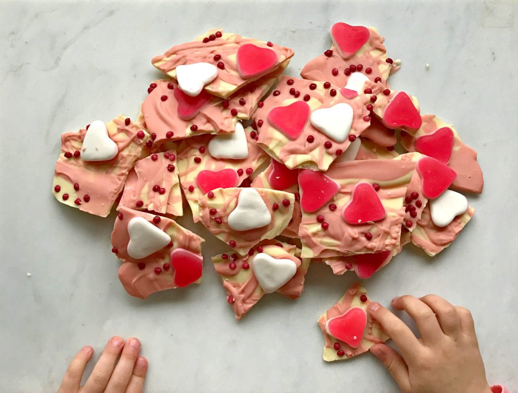 A childs hand holding a piece of chocolate bark with hearts and sprinkles on it for Valentine's Day. There is a big pile of bark next to her.