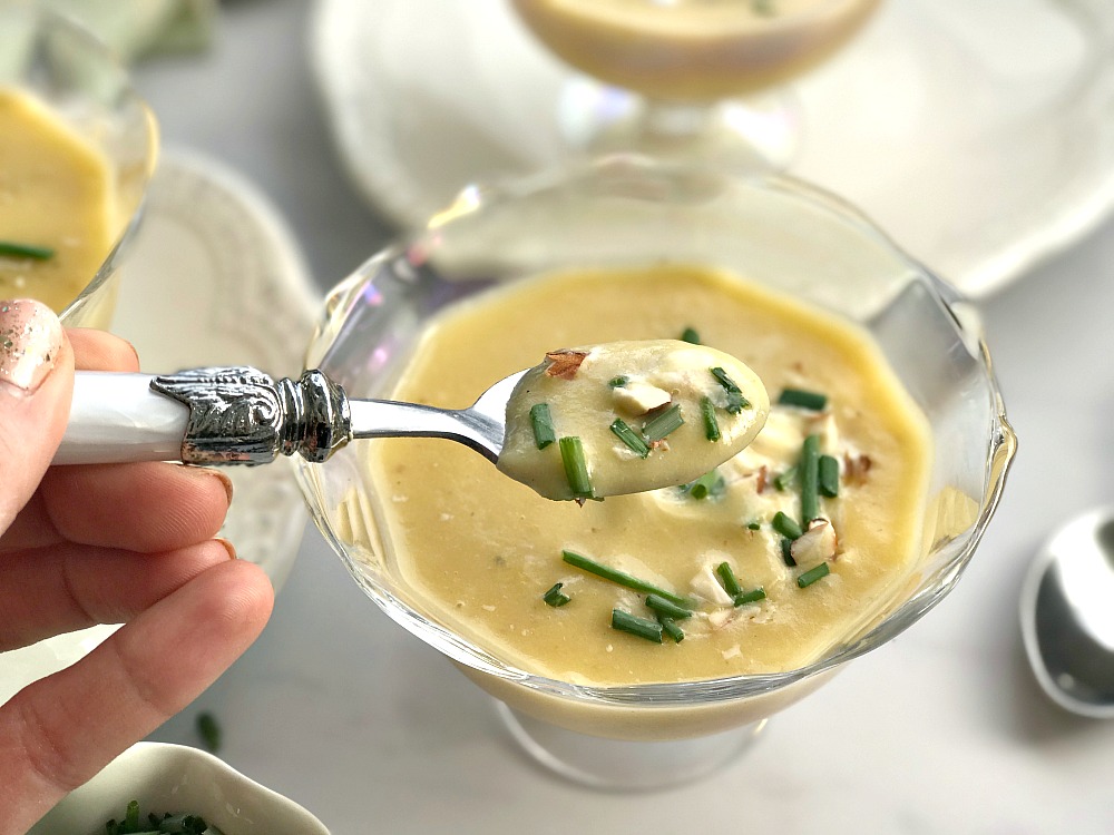 a hand holding a spoon with soup on it. The soup is in a glass bowl on the table.
