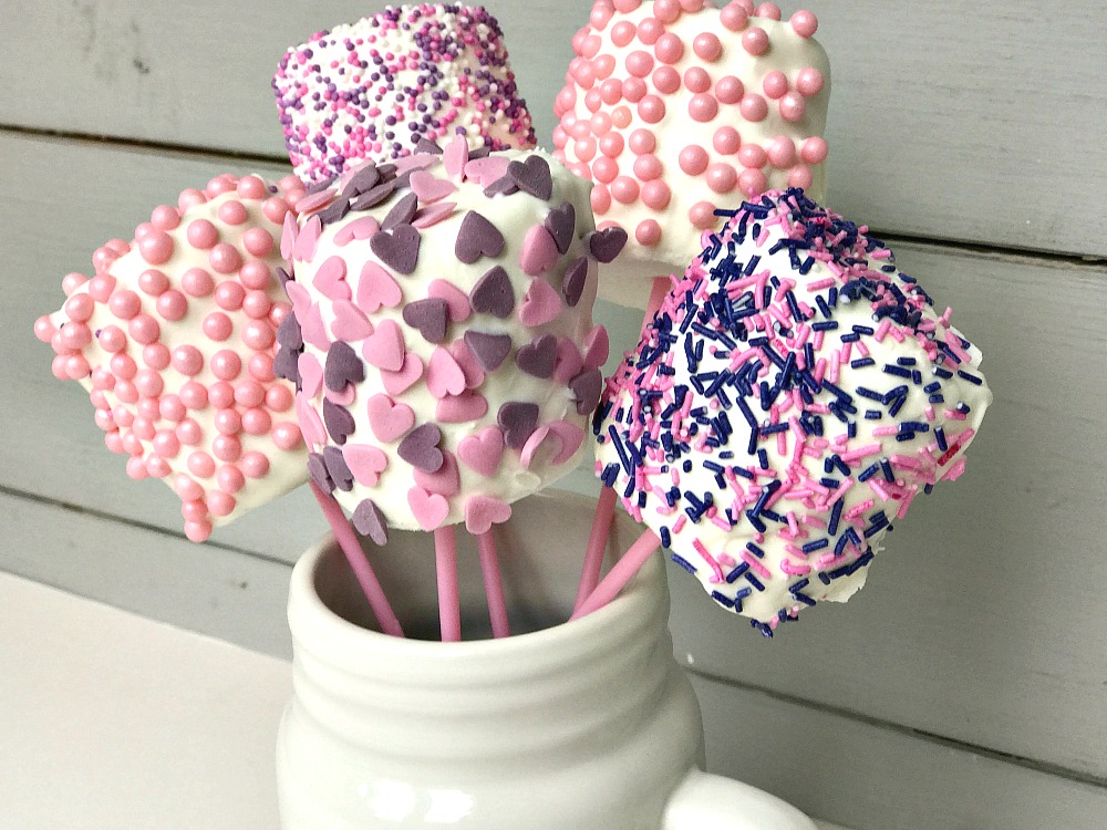 A white vase or mug with 5 marshmallow pops in it. They are on pink lolly sticks and covered with pink and purple hearts and sprinkles. Next to the base is 3 further marshmallow pops.