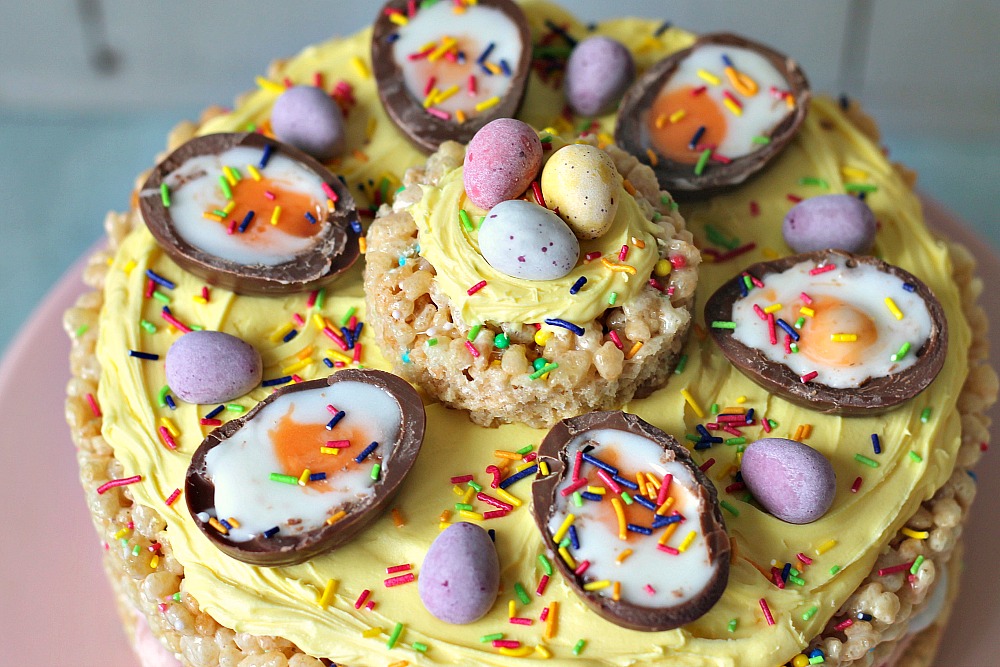 An Easter cake make out of rice crispy treats sits on a pink cake stand and topped with yellow icing, creme eggs and mini eggs