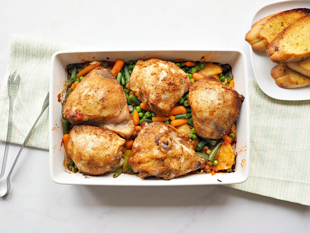 A white casserole dish with chicken and mixed vegetables in it. There is a dish of garlic bread on the outskirts of the photo.