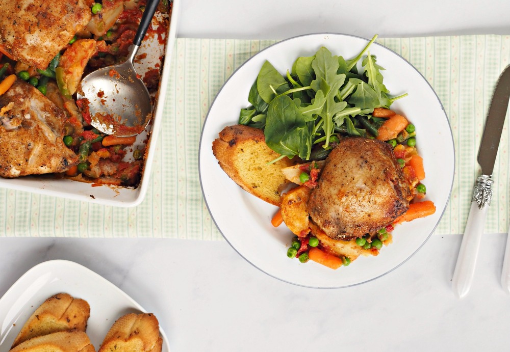 A plate with Rustic Chicken Casserole, mixed venetables, garlic bread and a leaf salad. Next to it is a large white dish where the meal came out of.