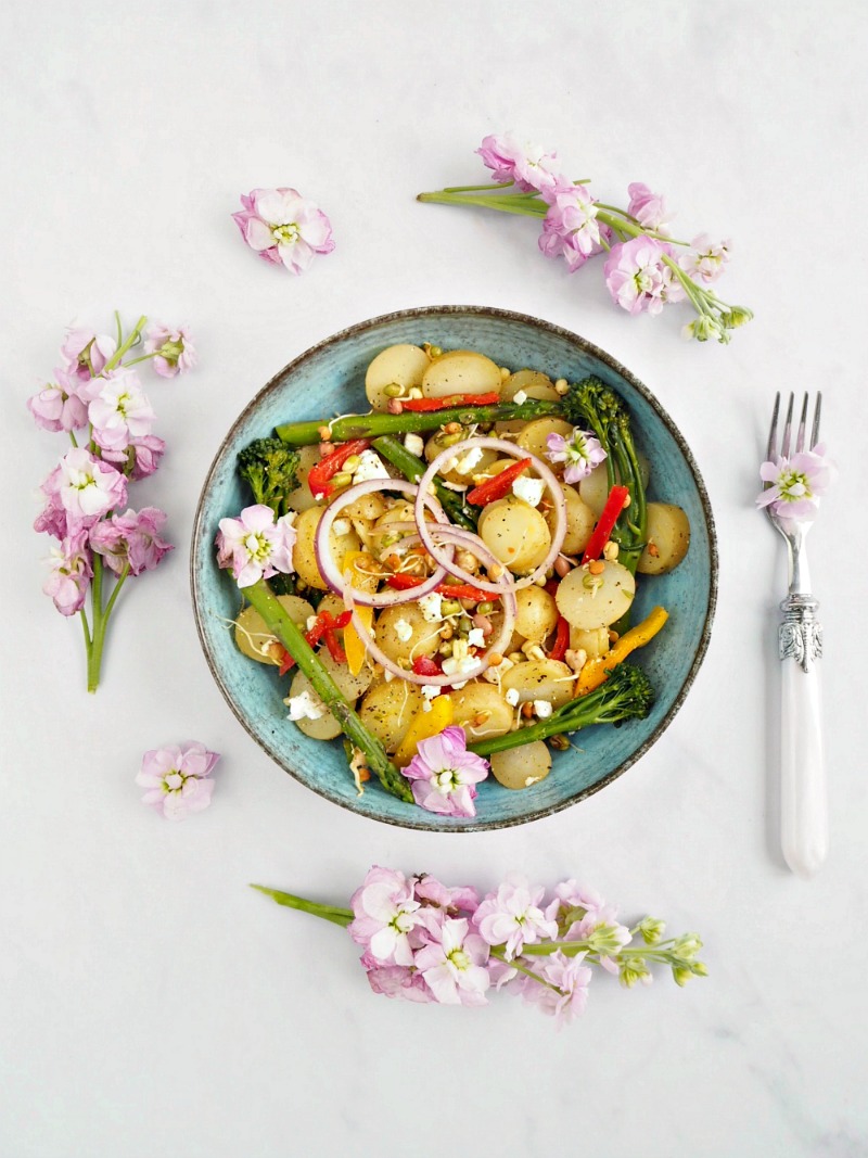 A bowl of mango, Lime and Chilli Potatoes with onion, peppers and tenderstem broccoli. The bowl and surrounding area is decorated with pink flowers.