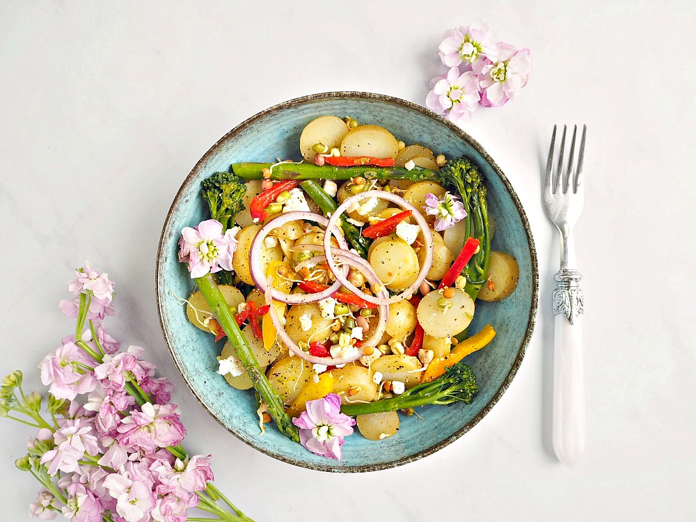 A bowl of mango, Lime and Chilli Potatoes with onion, peppers and tenderstem broccoli. The bowl and surrounding area is decorated with pink flowers.