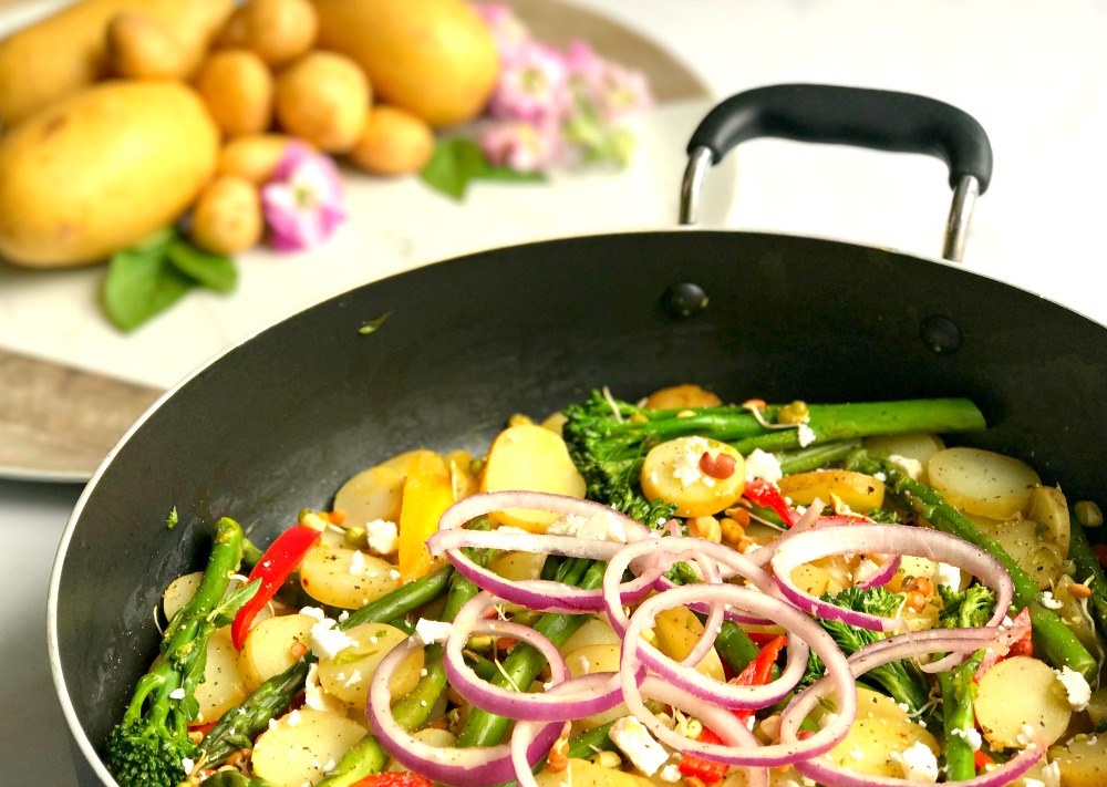 A frying pan containing Lime and Chilli Potatoes with onion, peppers and tenderstem broccoli. In the background is a pile of potatoes and pink flowers.