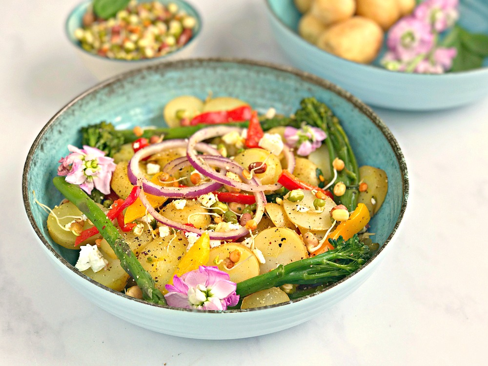 There are 3 blue bowls. One contains mango, Lime and Chilli Potatoes with onion, peppers and tenderstem broccoli. Another contains lentil sprout mix. The last holds potatoes and some pink flowers.