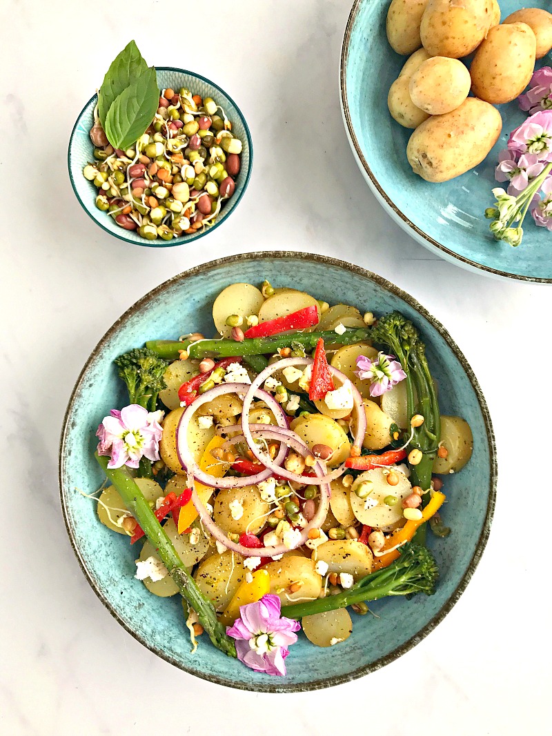 There are 3 blue bowls. One contains mango, Lime and Chilli Potatoes with onion, peppers and tenderstem broccoli. Another contains lentil sprout mix. The last holds potatoes and some pink flowers.