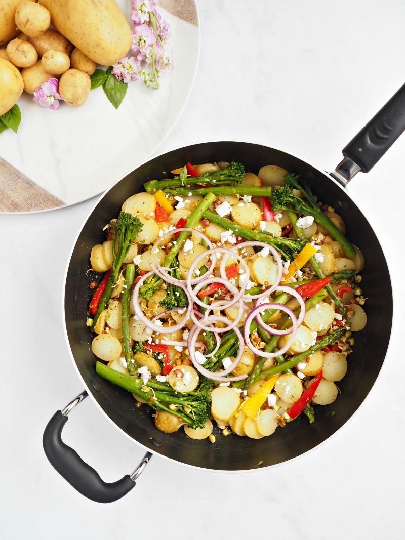 A frying pan containing Lime and Chilli Potatoes with onion, peppers and tenderstem broccoli. A plate is next to it with a pile of potatoes and pink flowers.