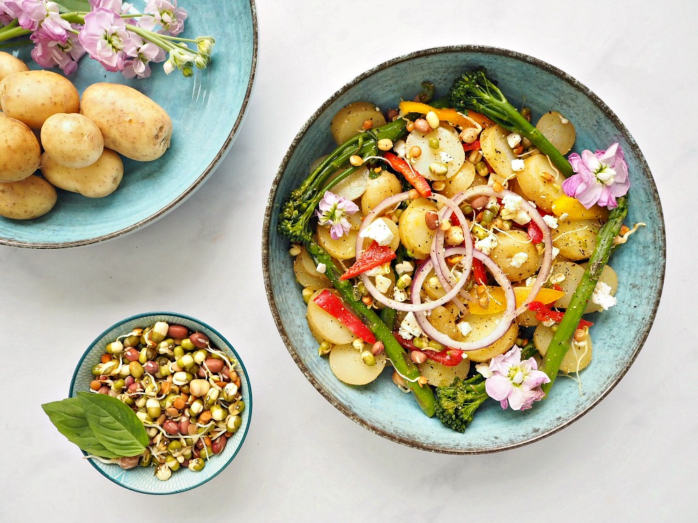 There are 3 blue bowls. One contains mango, Lime and Chilli Potatoes with onion, peppers and tenderstem broccoli. Another contains lentil sprout mix. The last holds potatoes and some pink flowers.