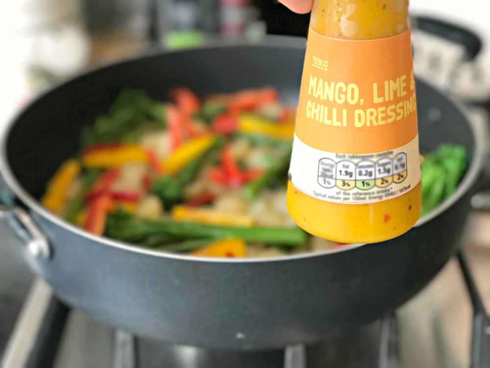 A close up on a bottle of Mango, lime and chilli dressing from Tesco, with a frying pan of vegetables in the background.