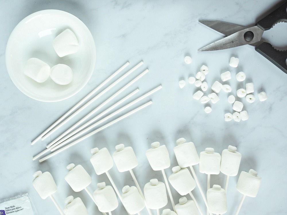 White giant marshmallow, mini marshmallows, lolly sticks and scissors lie on a counter, with the start of lego marshmallow pops below