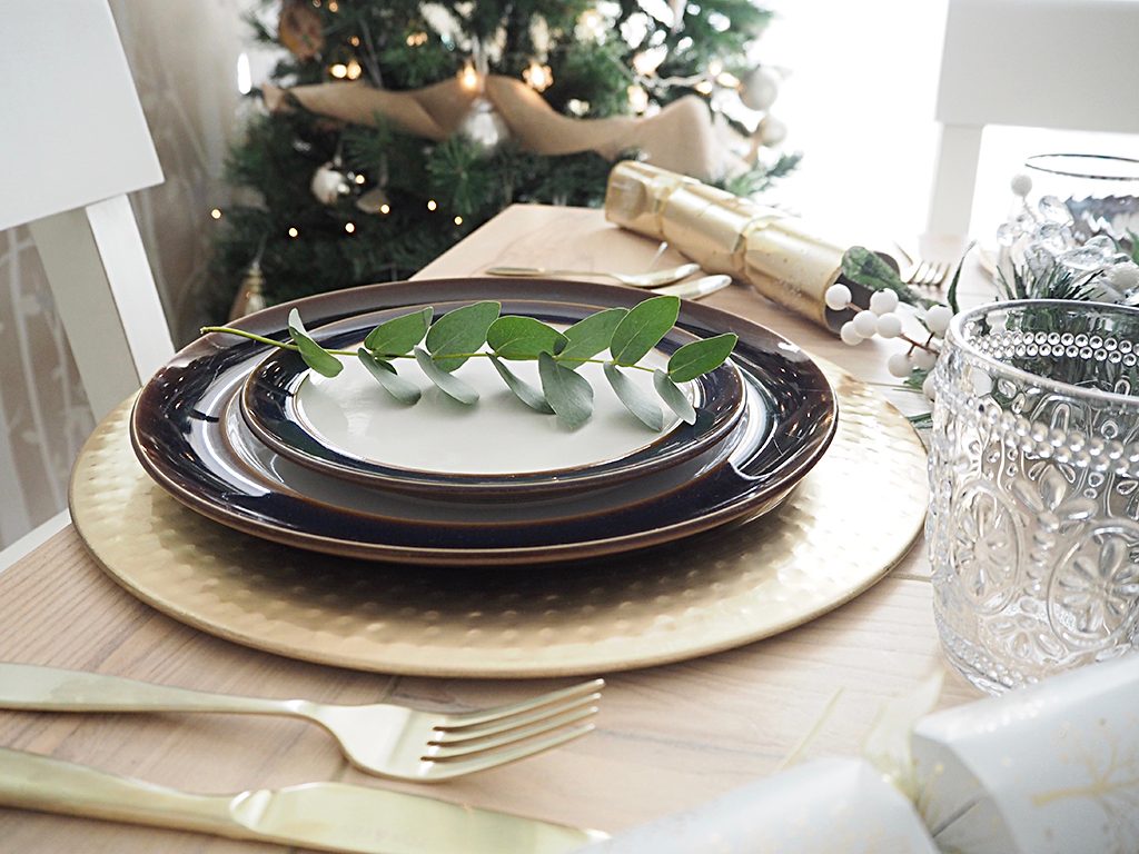 A table place setting for Christmas. A gold charger with blue white and gold crockery and gold cutlery, white and gold crackers and some greenery