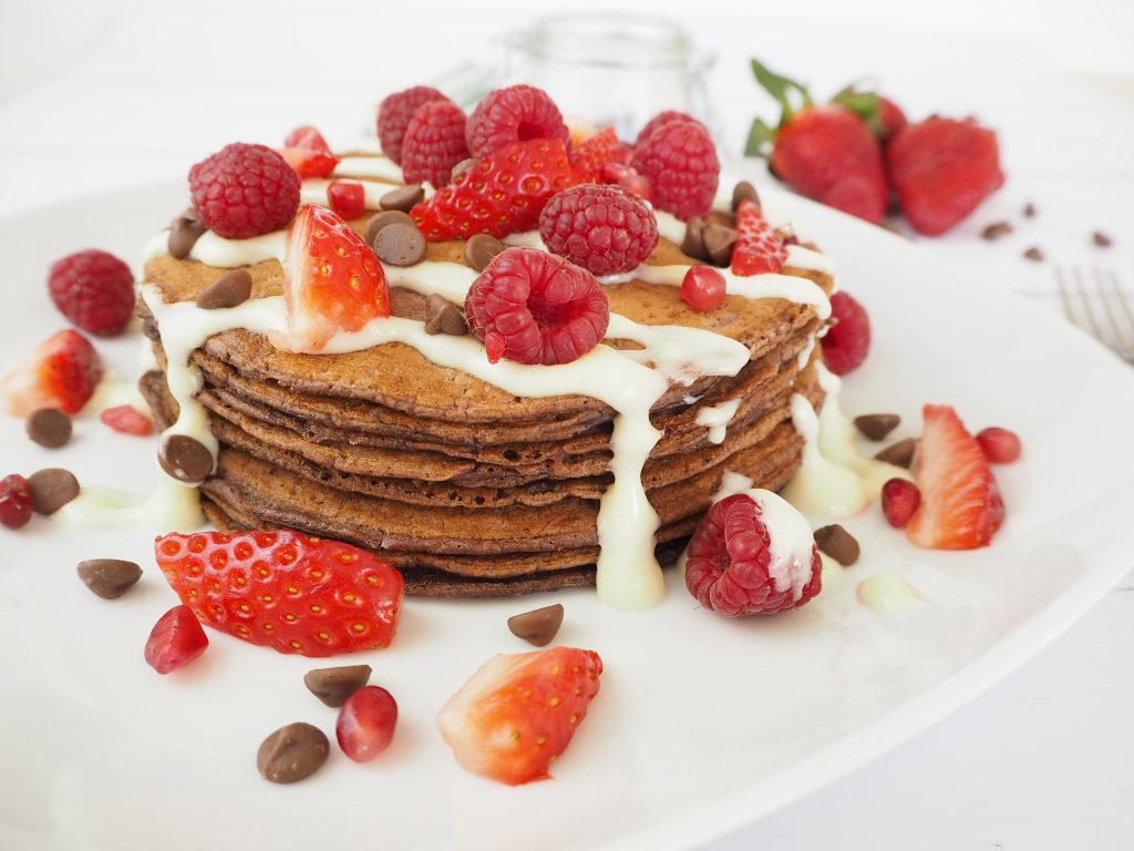 A stack of chocolate pancakes, topped with a cream cheese glaze, strawberries, raspberries and chocolate chips