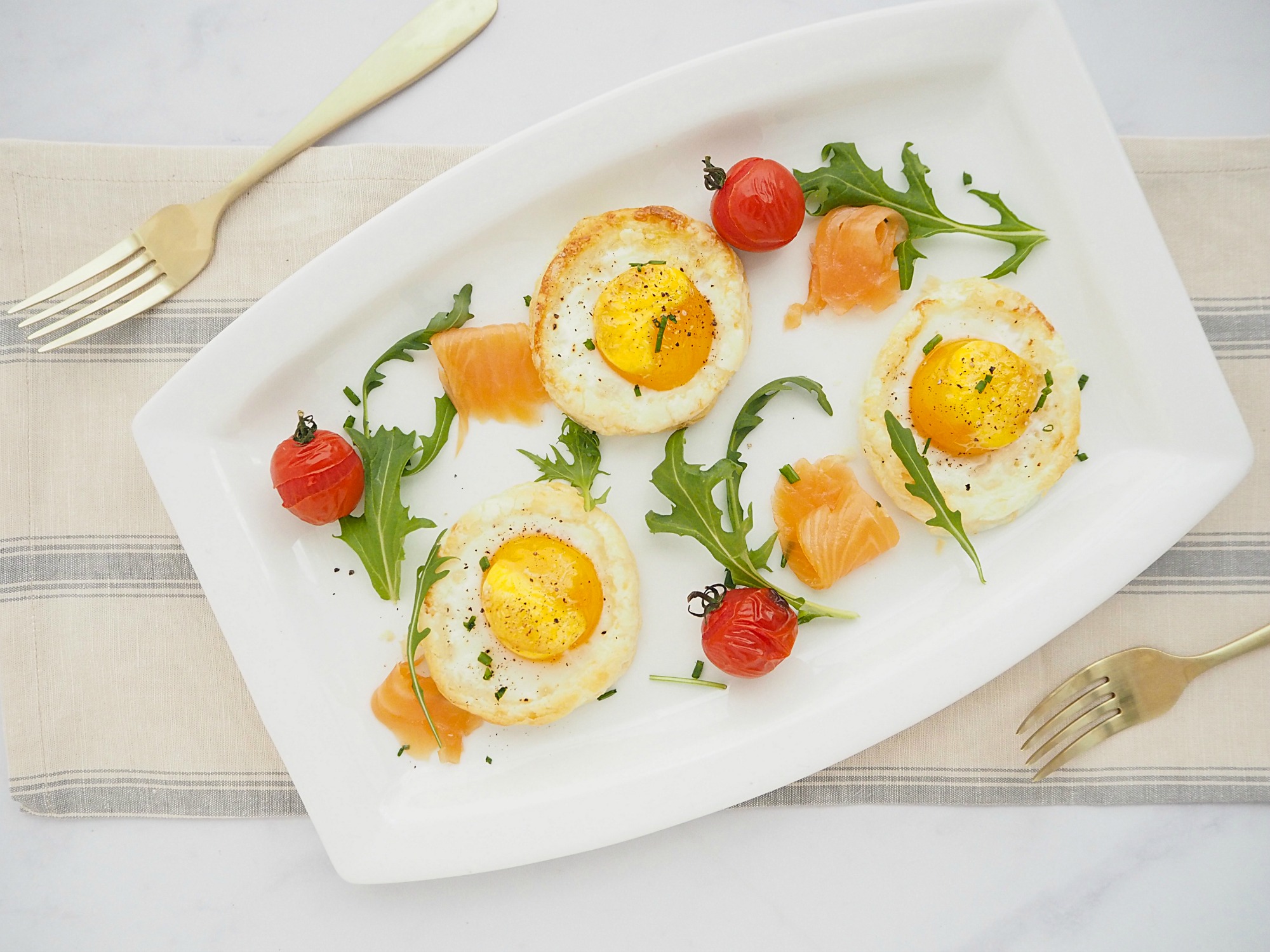 A white rectangular plate with 3 baked eggs, smoked salmon, rocket and roasted cherry tomatoes