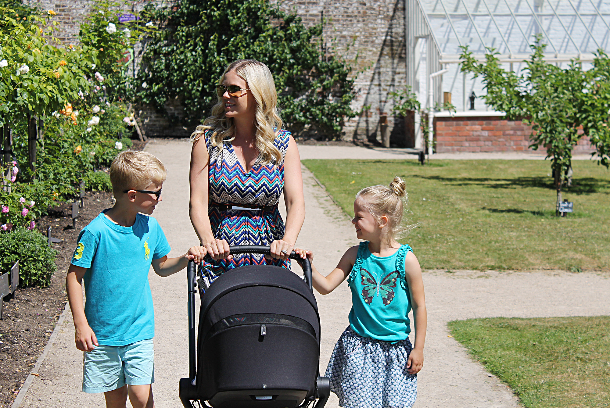 A woman wearing a patterned dress is walking in a park with 2 children walking beside her and pushing a black pram