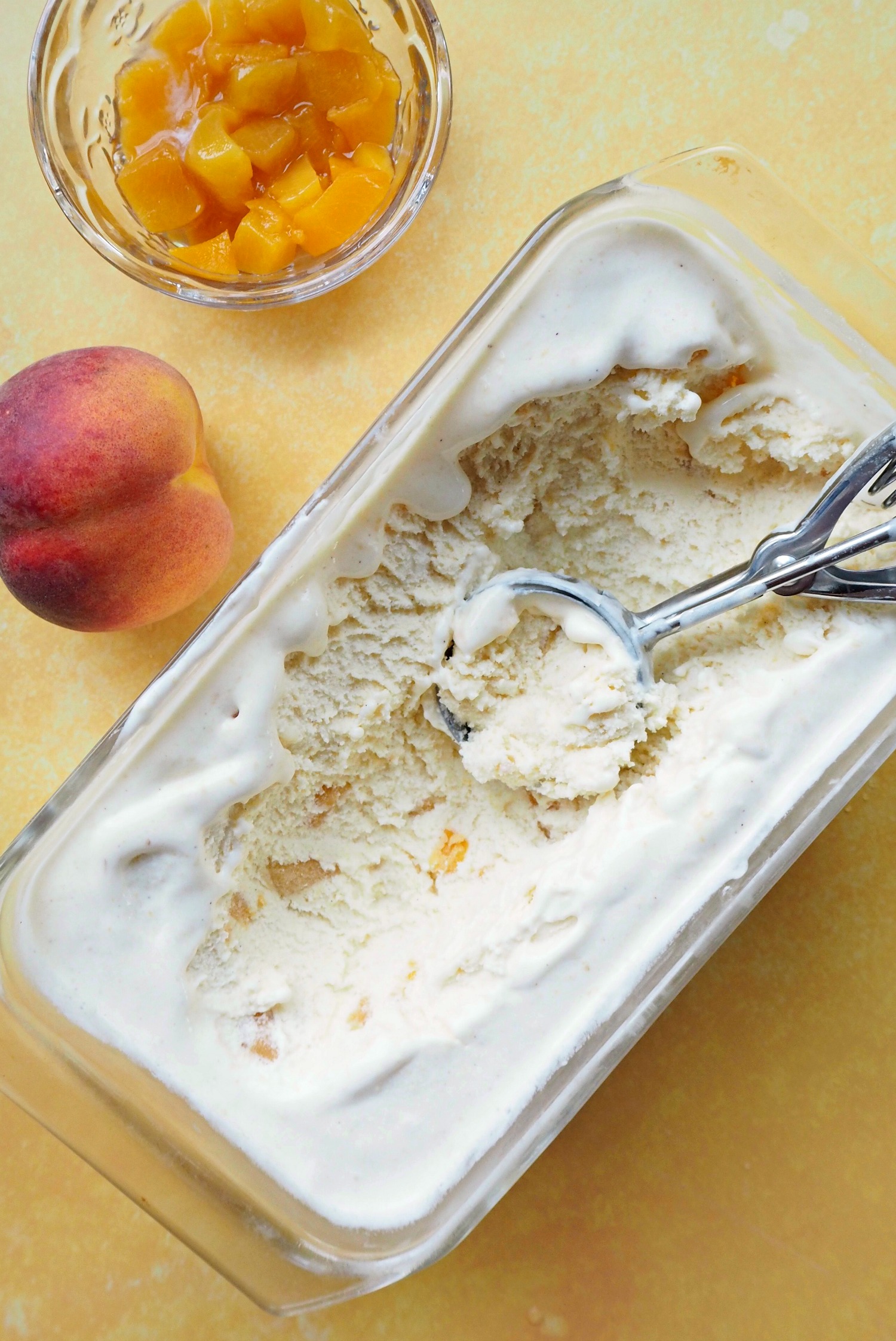 A tub of ice cream with an ice cream scoop in it, next to a dish of peach chunks and a whole peach.