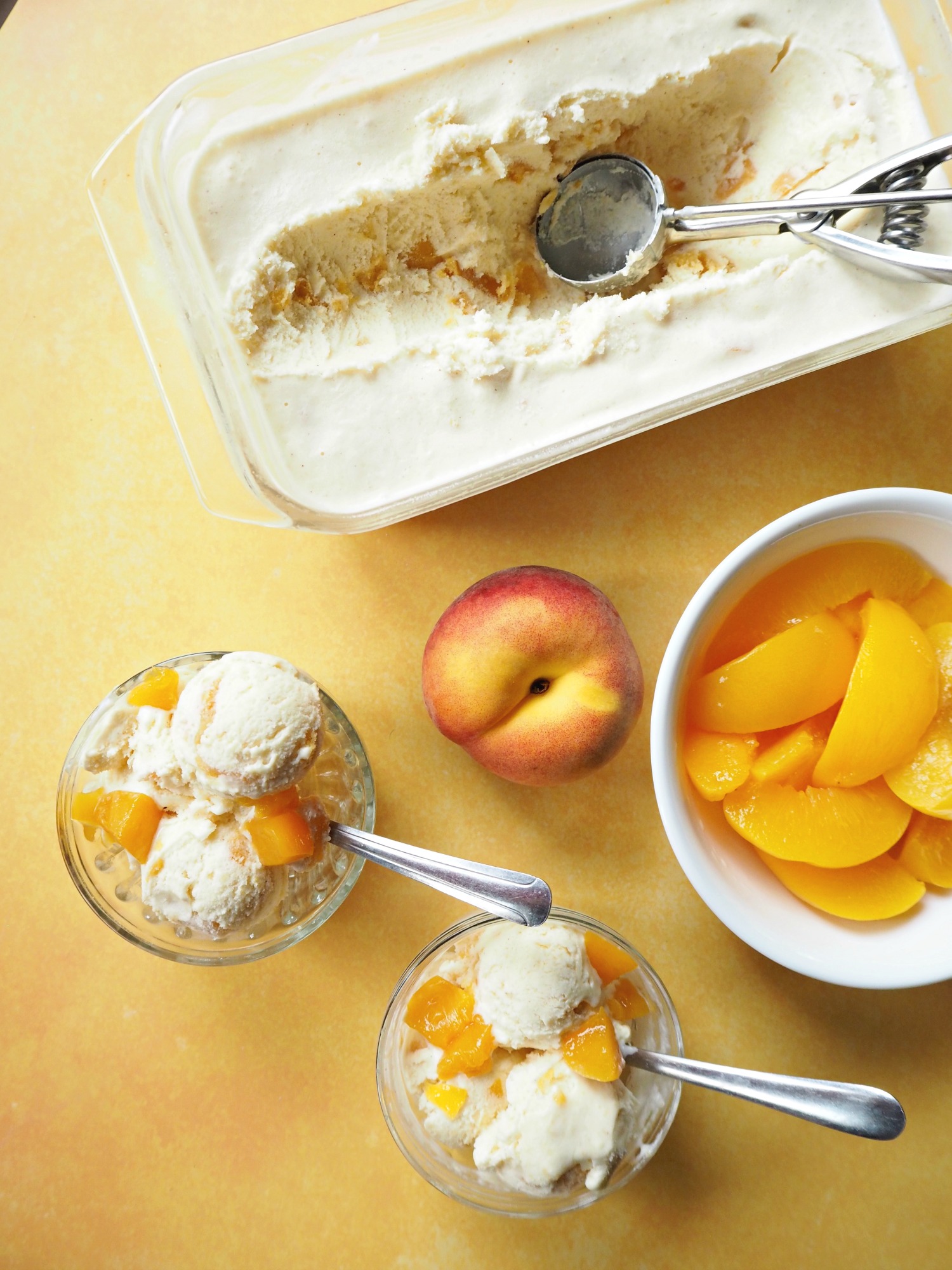 A tub of homemade ice cream with an ice cream scoop in it, next to a couple of bowls of peach ice cream, a whole peach and a bowl of peach slices.
