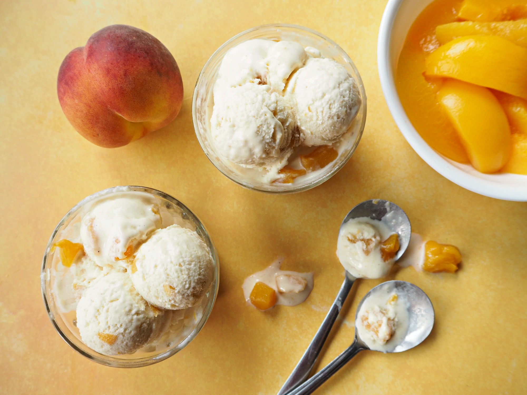 A top down view of 2 glass bowls filled with peach ice cream, 2 dirty spoons, a peach and a bowl of sliced peaches.
