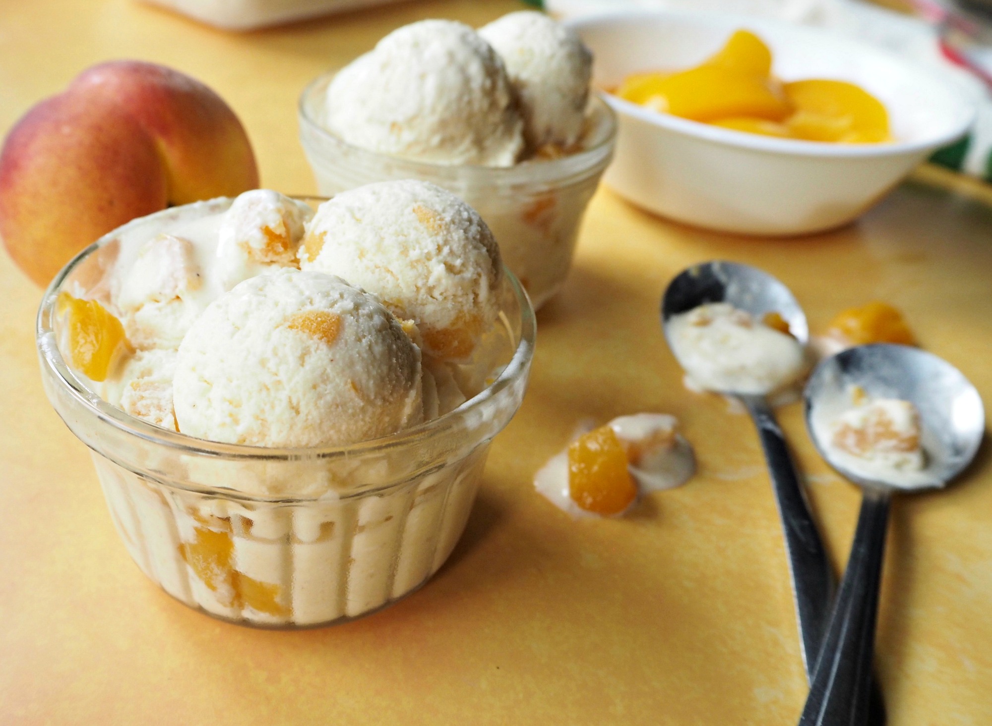 Bowls filled with schools of peach ice cream. A whole peace and a bowl of sliced peaches in the background, along with 2 spoons with ice cream on them.