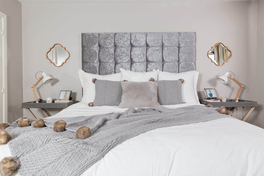 A bed with a large grey headboard. It has white sheets on it and a set of grey pillows and a grey throw diagonally across it