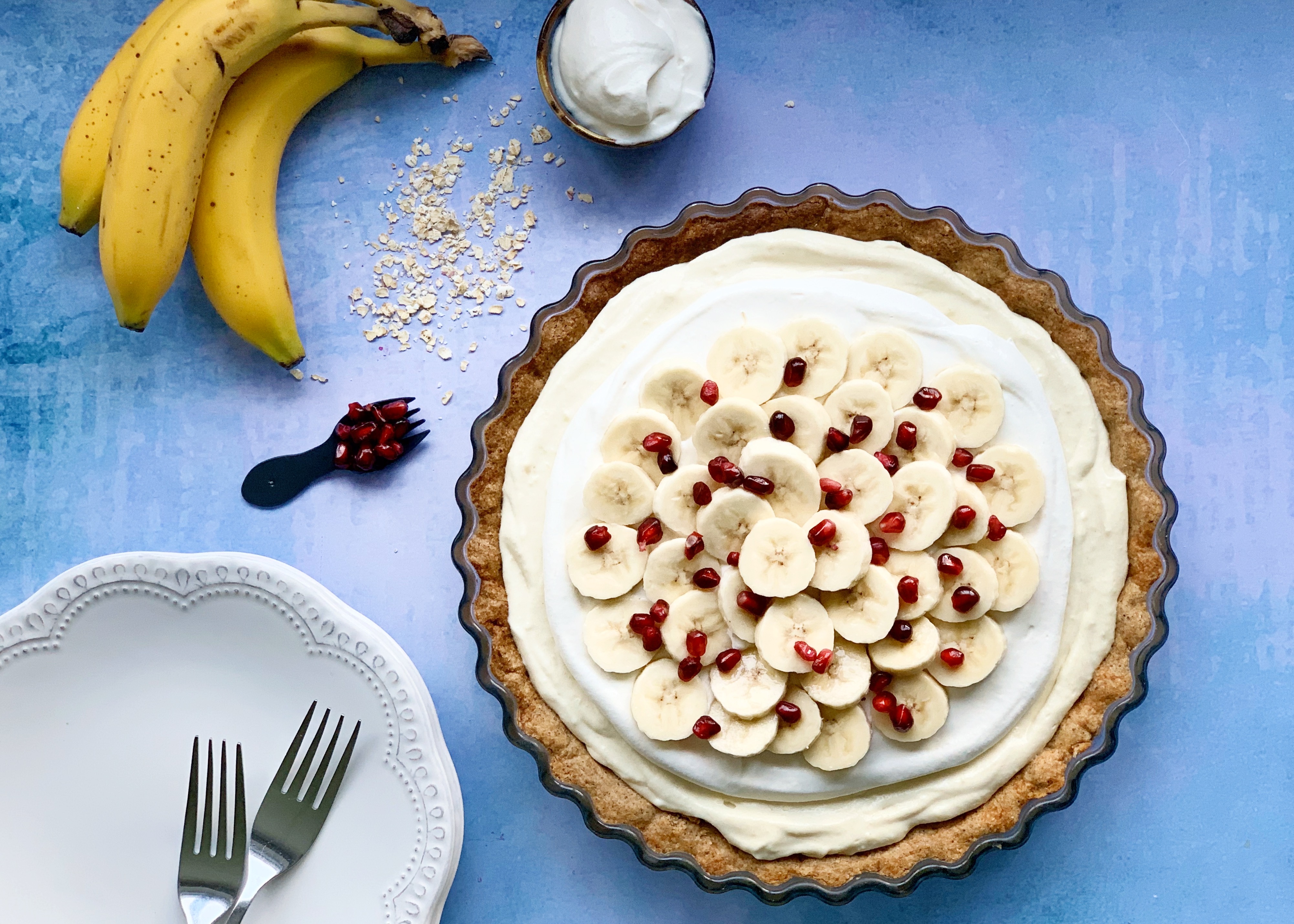 Vegan Banana Cream Pie with Pomegranate next to a plate with 2 forks on it, a bunch of bananas, some oats, cream and extra pomegranate. They are all sitting on a blue background.