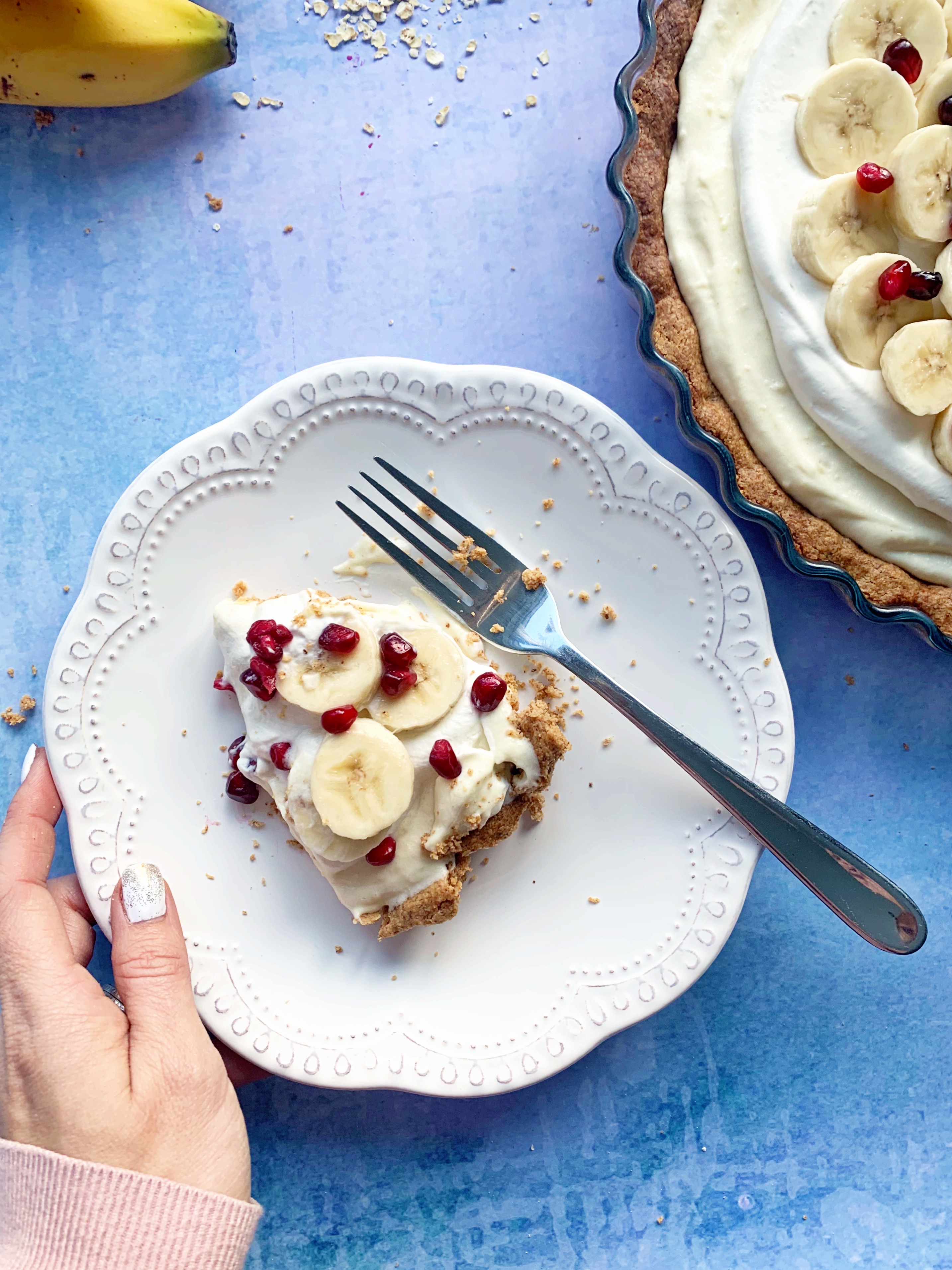 A slice of Vegan Banana Cream Pie with Pomegranate on a white plate with a fork. The rest of the pie is partially visable to the right of the plate.