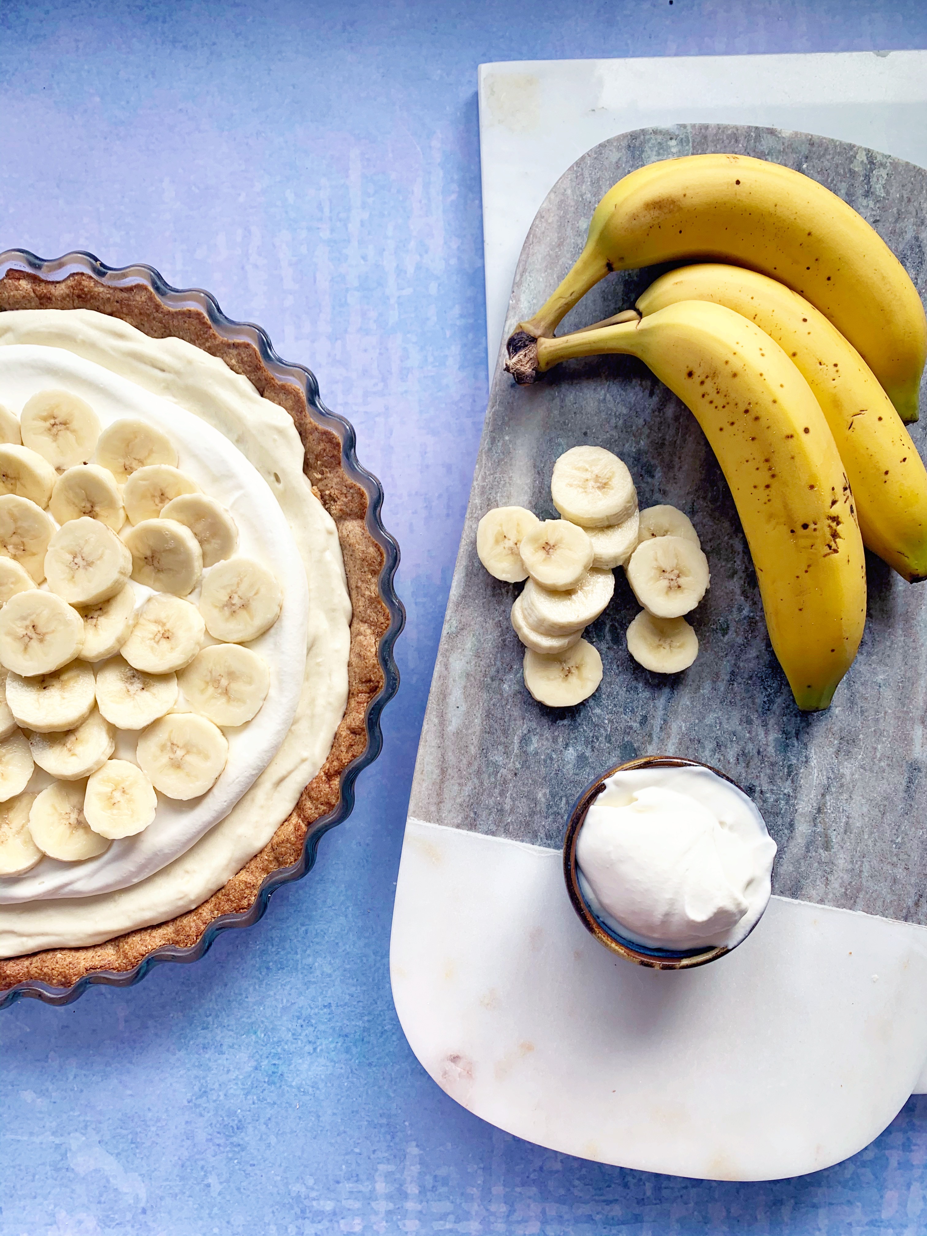 A vegan banana vream pie sits next to some extra sliced banana, 3 whole bananas and some coconut cream