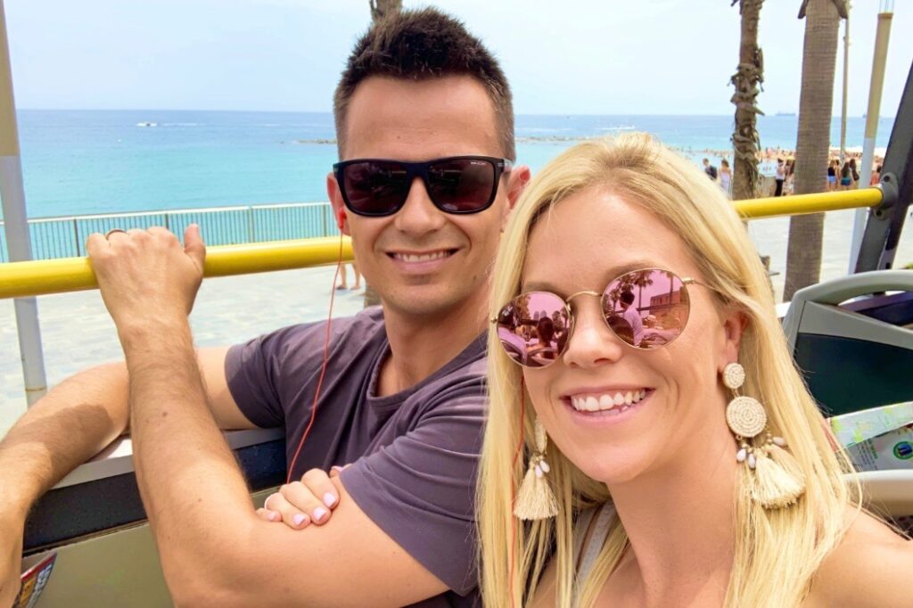 A man and woman wearing sunglasses as they sit on the top deck of an open top bus. Behind them is the ocean