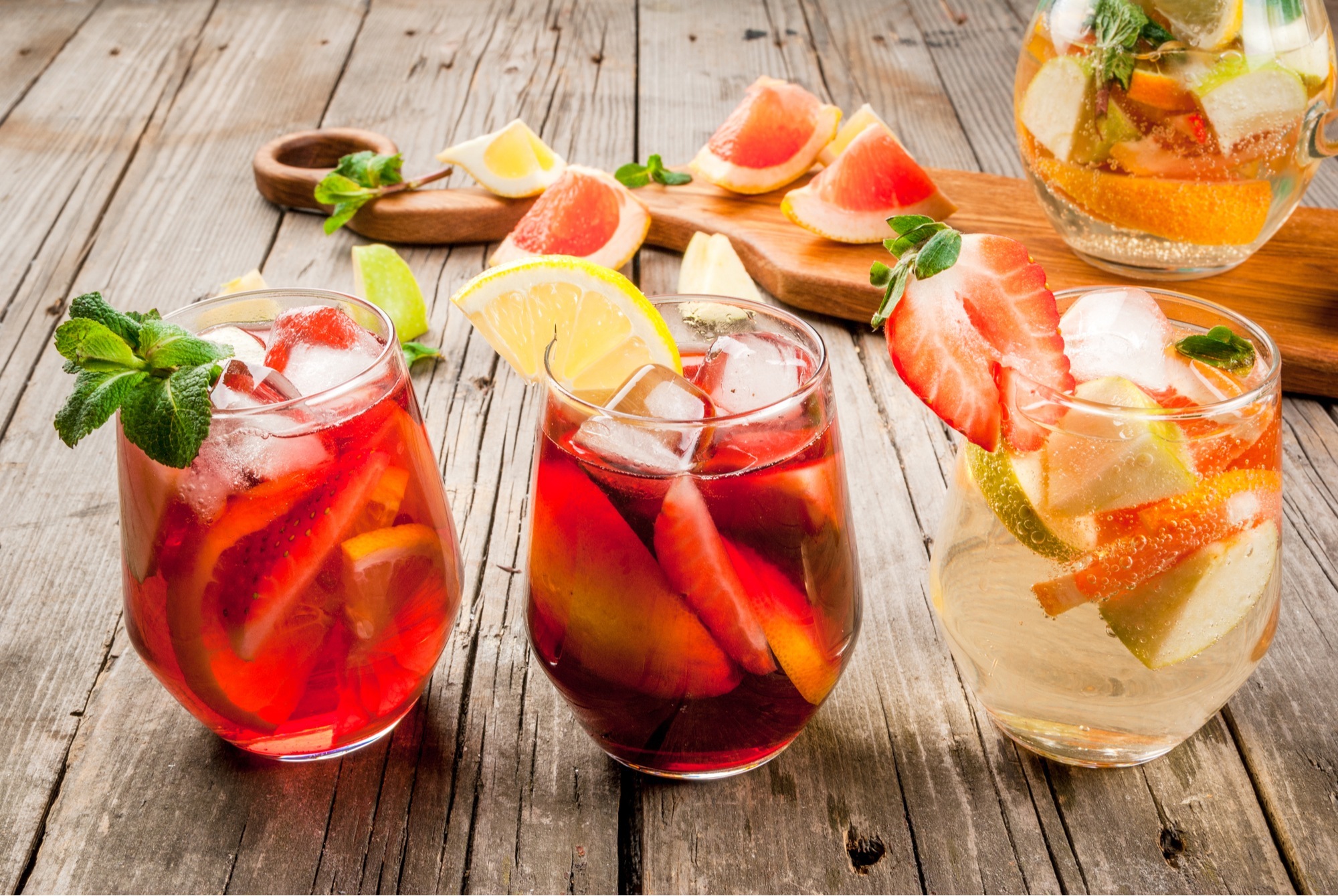 3 cocktails in short glasses sat on a wooden table. The cocktail glasses are full of fruit and garnished with fruit.