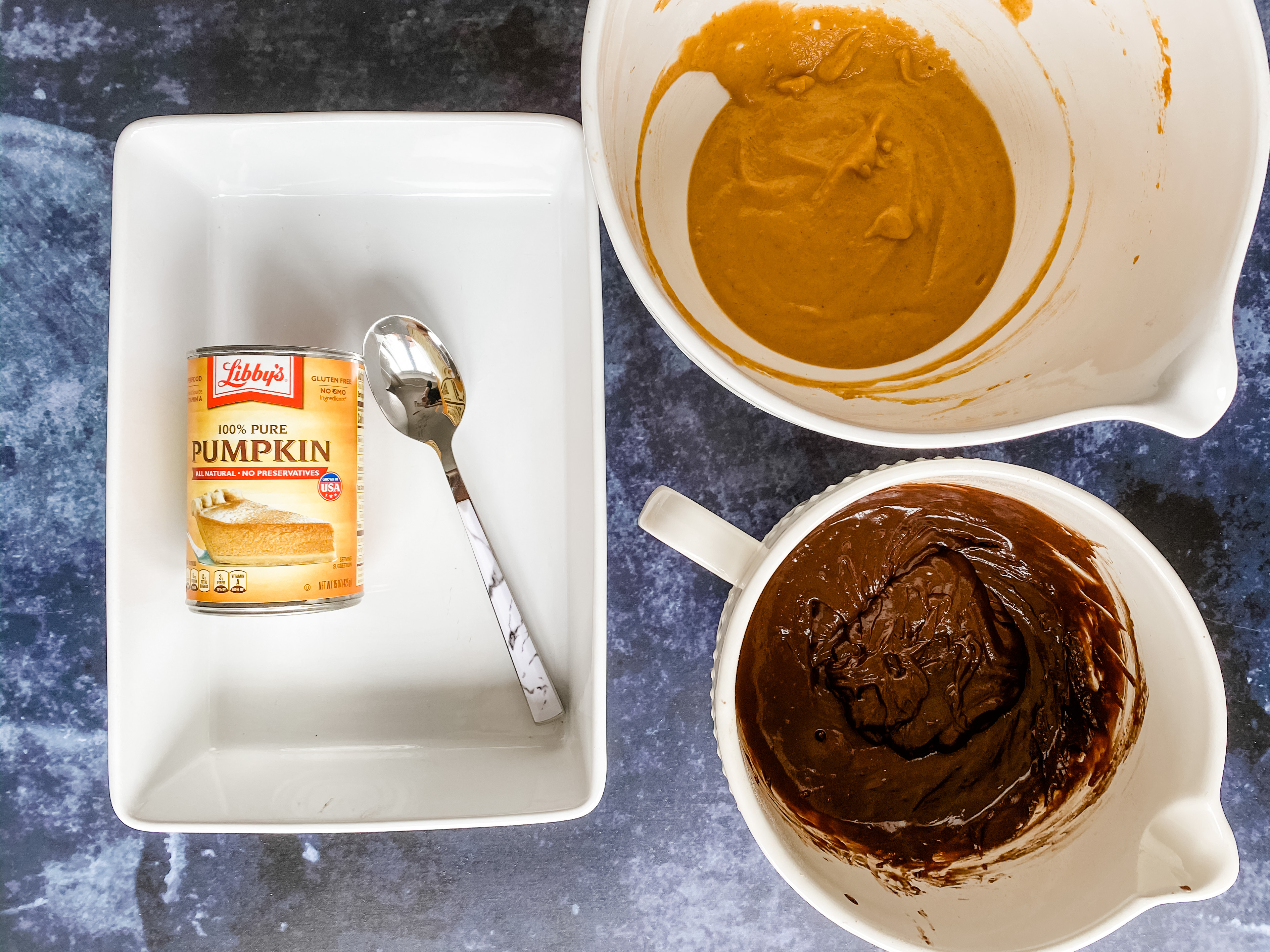 A white rectangular dish with a can pf pumpkin puree and a spoon in it. A round white bowl with pumpkin puree in it and a white mixing jug with brownie mix in it