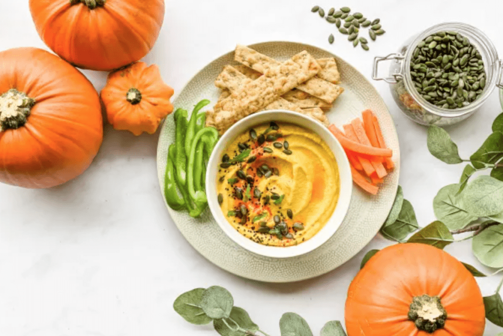 A bowl of pumpkin hummus on a plate with crudities and surrounded by mini pumpkins