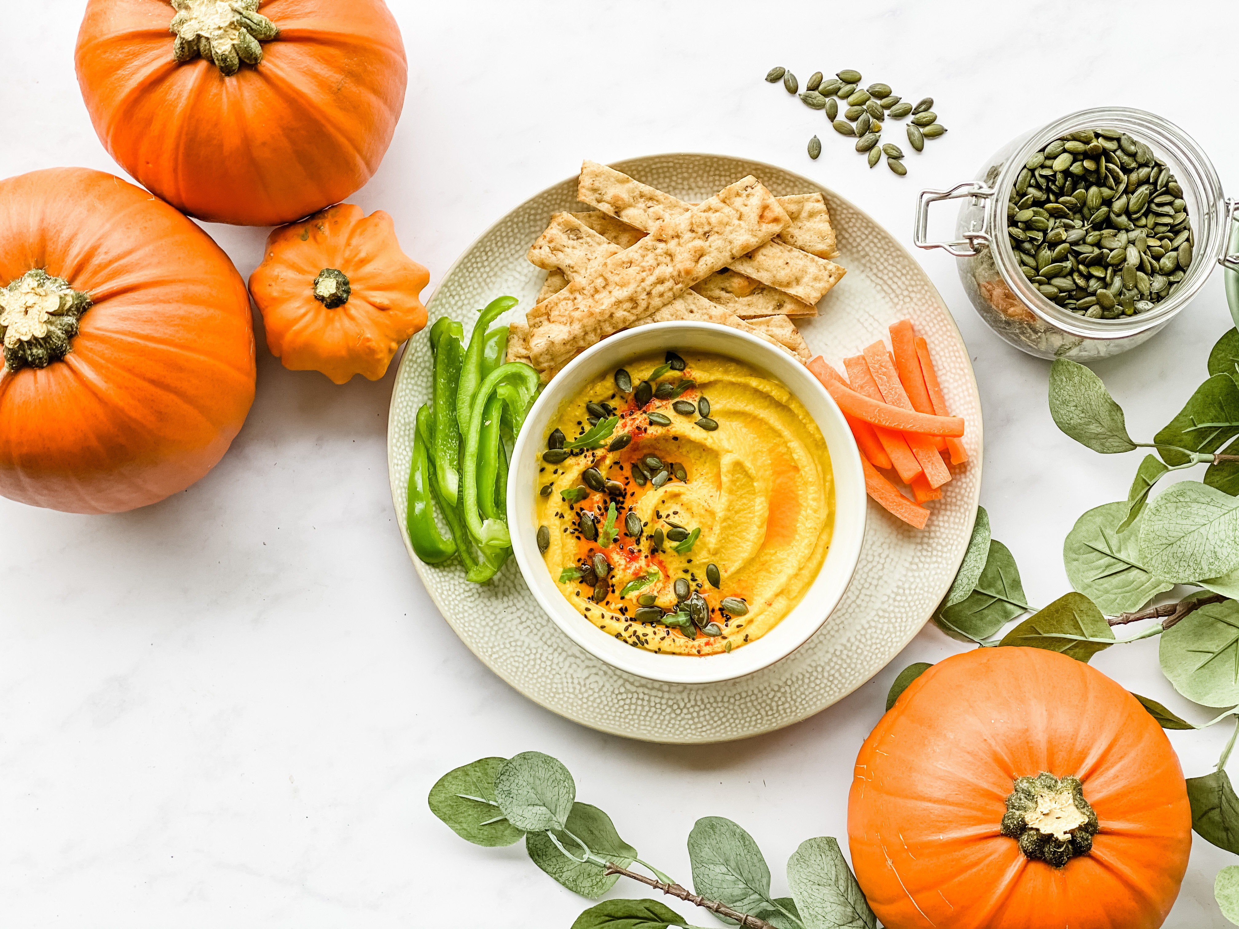 A white bowl set on a plate. The bowl has orange coloured pumpkin hummus topped with chilli oil and pumpkin seeds. On the plate is some sliced carrot and peppers and some strips of bread. Surrounding the plate is fall decor in the form of pumpkins and leaves.