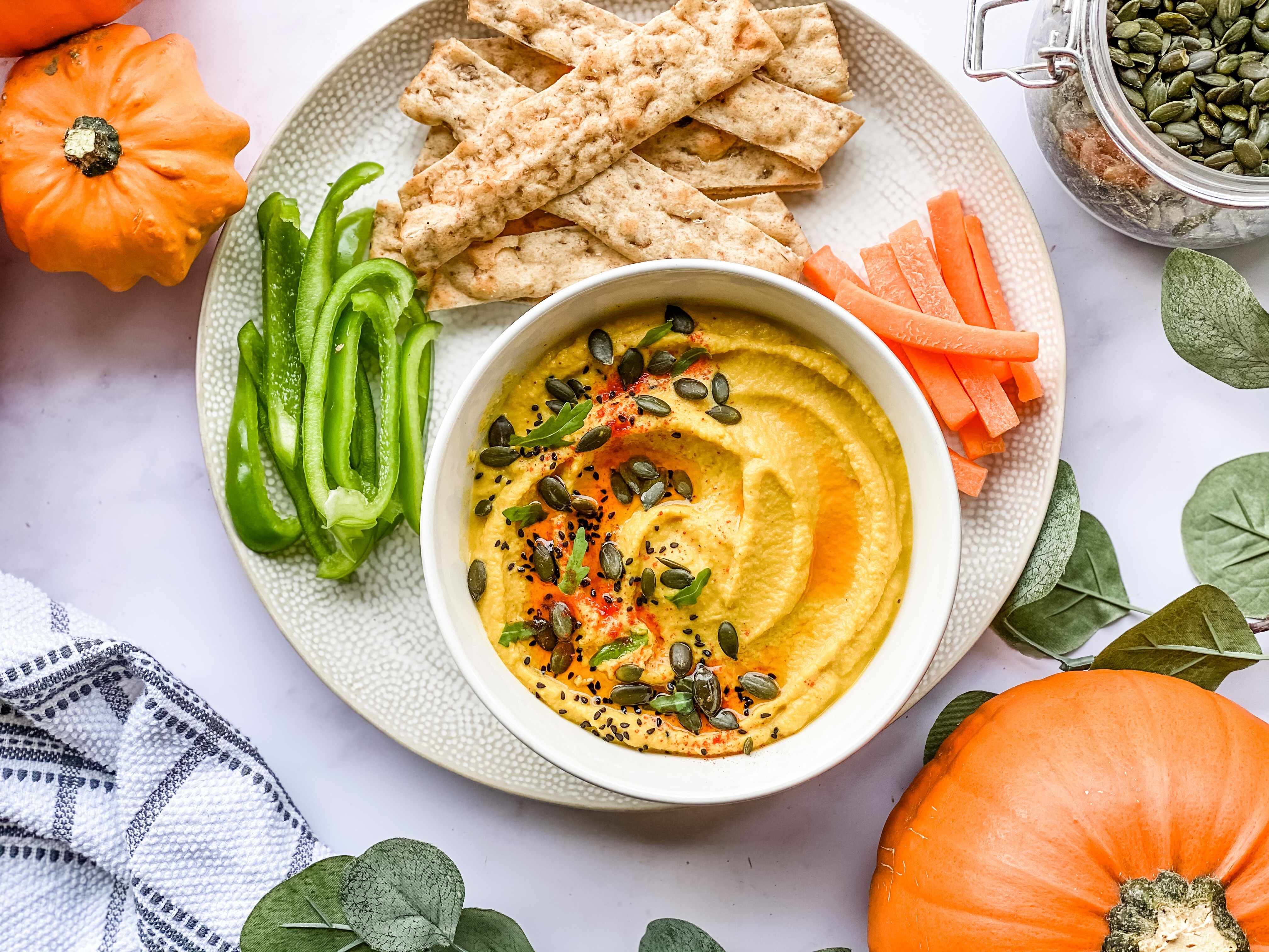 A white bowl set on a plate. The bowl has orange coloured pumpkin hummus topped with chilli oil and pumpkin seeds. On the plate is some sliced carrot and peppers and some strips of bread. Surrounding the plate is fall decor in the form of pumpkins and leaves.