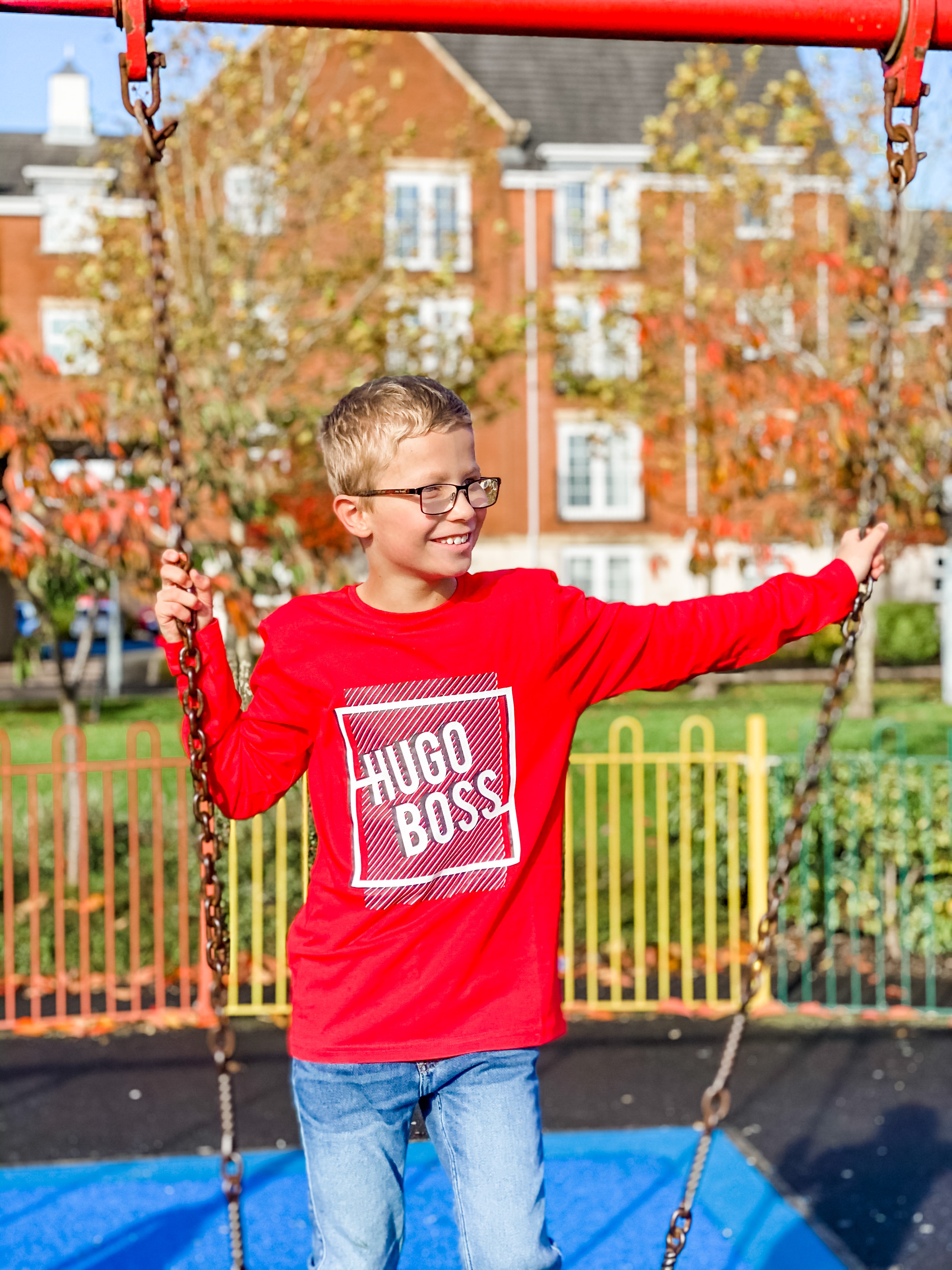 A pre-teen boy with blonde hair and glasses, wearing a red t-shirt, stands on a swing.