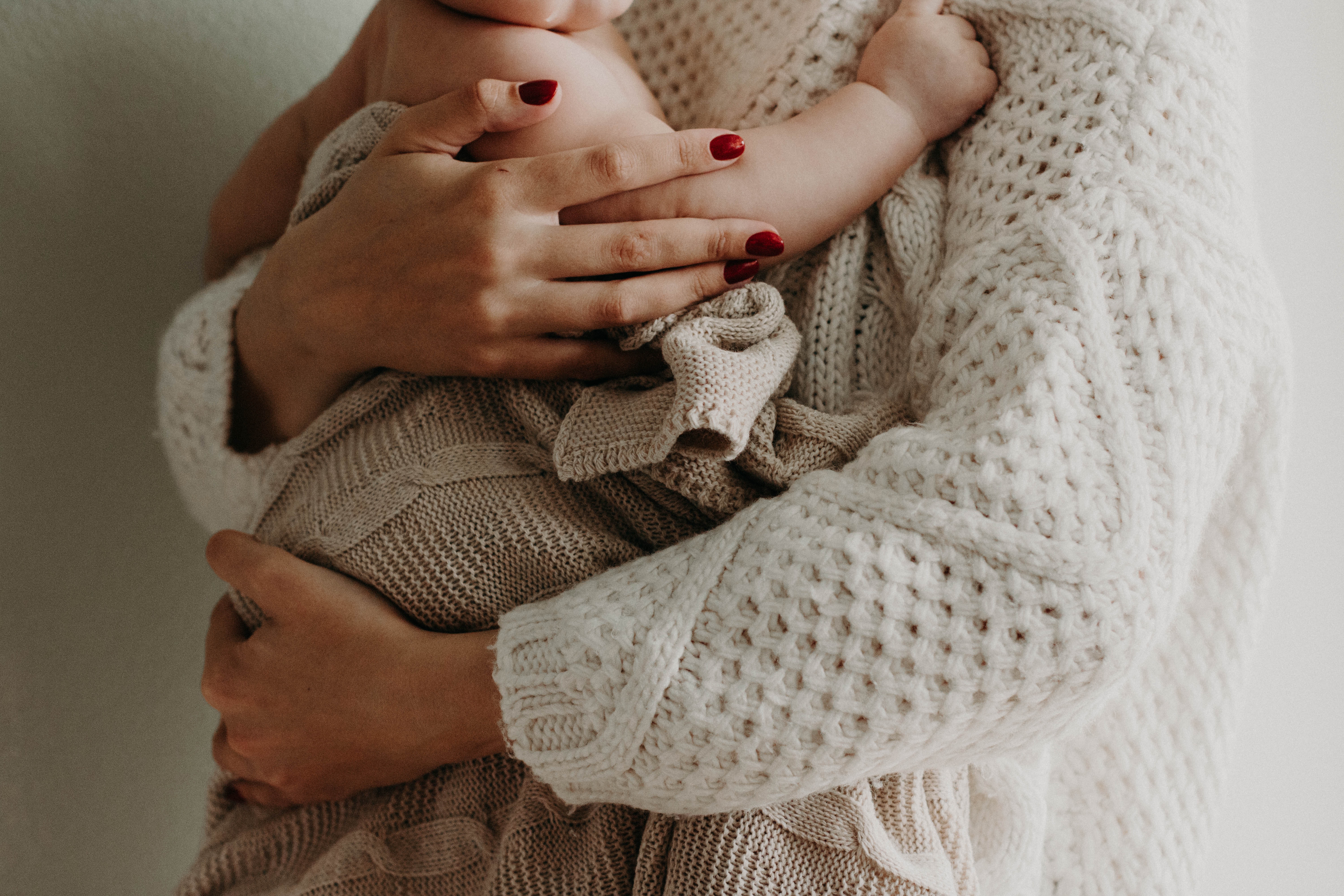 The torso of a woman holding a baby. The woman's jumper and the baby's blanket are in natural earthy colours