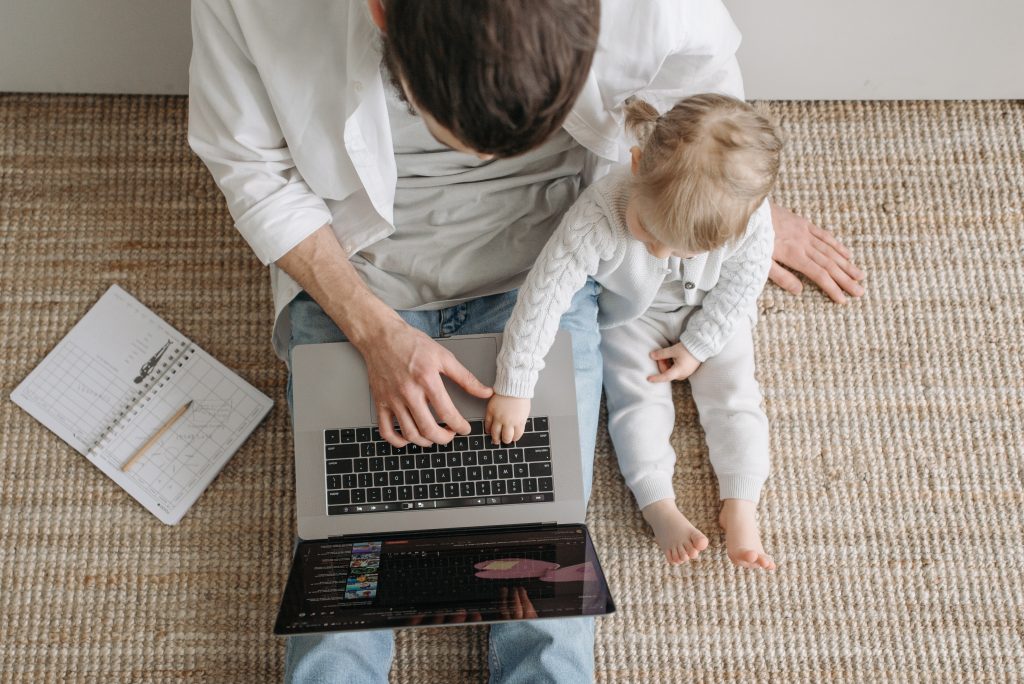 A man sits on the floor with his laptop. A baby sits with him and reaches over to touch the keyboard.