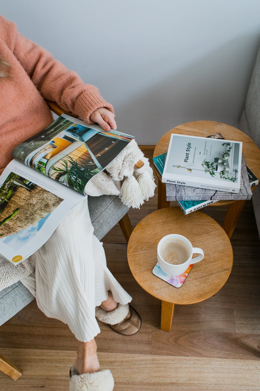 unrecognizable woman reading magazine in armchair near table with mug