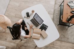 A bird's eye view of a woman sitting at a white desk. She is working on a laptop with notebooks around the desk