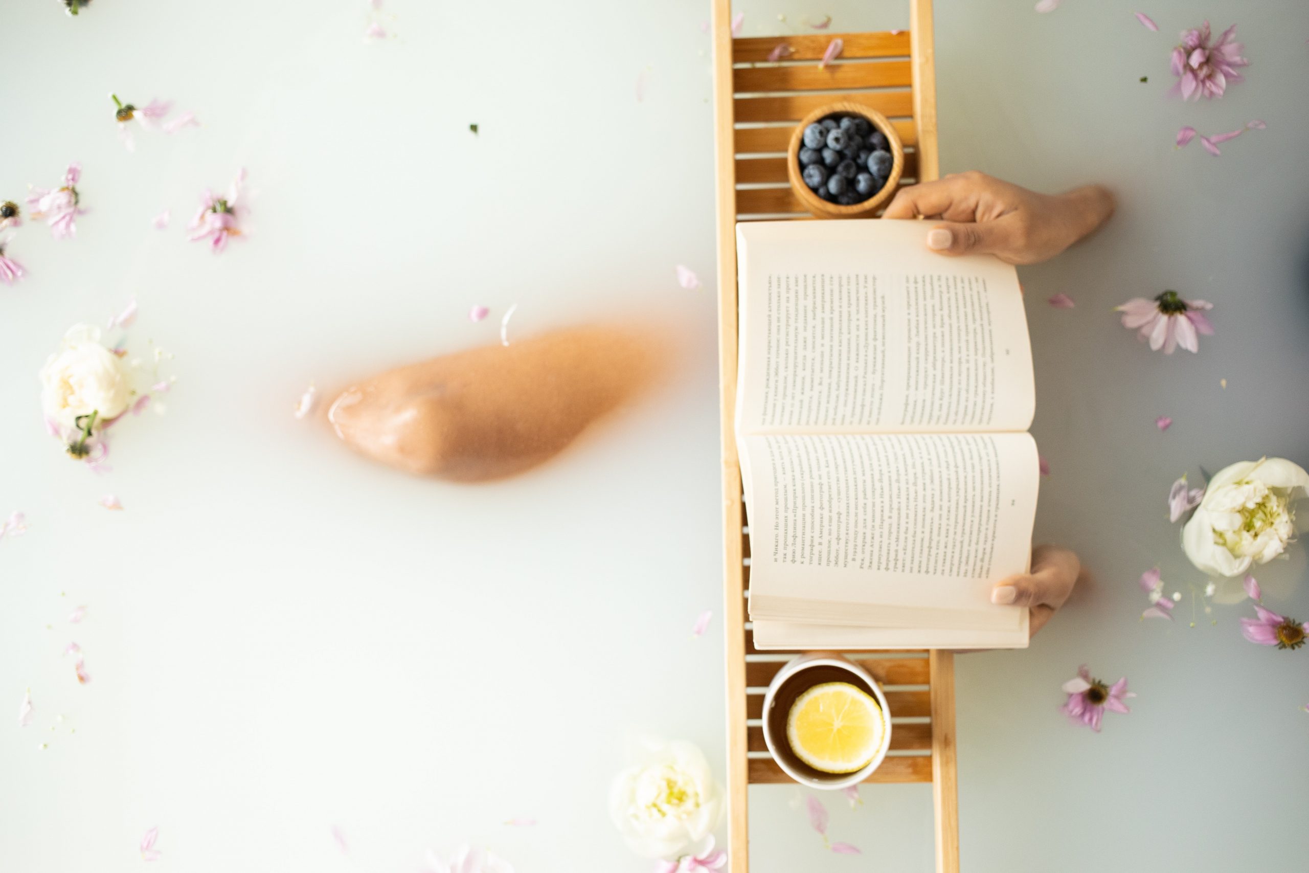 A milky bath with dried flowers in it. A person is in the water with their knee rising above the water level. They are reading a book, leaning on a wooden bath tray