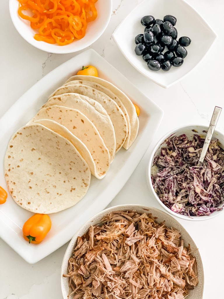 A top down view of some pulled pork, tortillas, red cabbage slaw, sliced peppers and olives