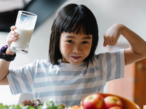 A young girl in a blue and white striped t-shirt. She is holding a glass of milk and has milk around her mouth, and has riased her arms up to show her muscles