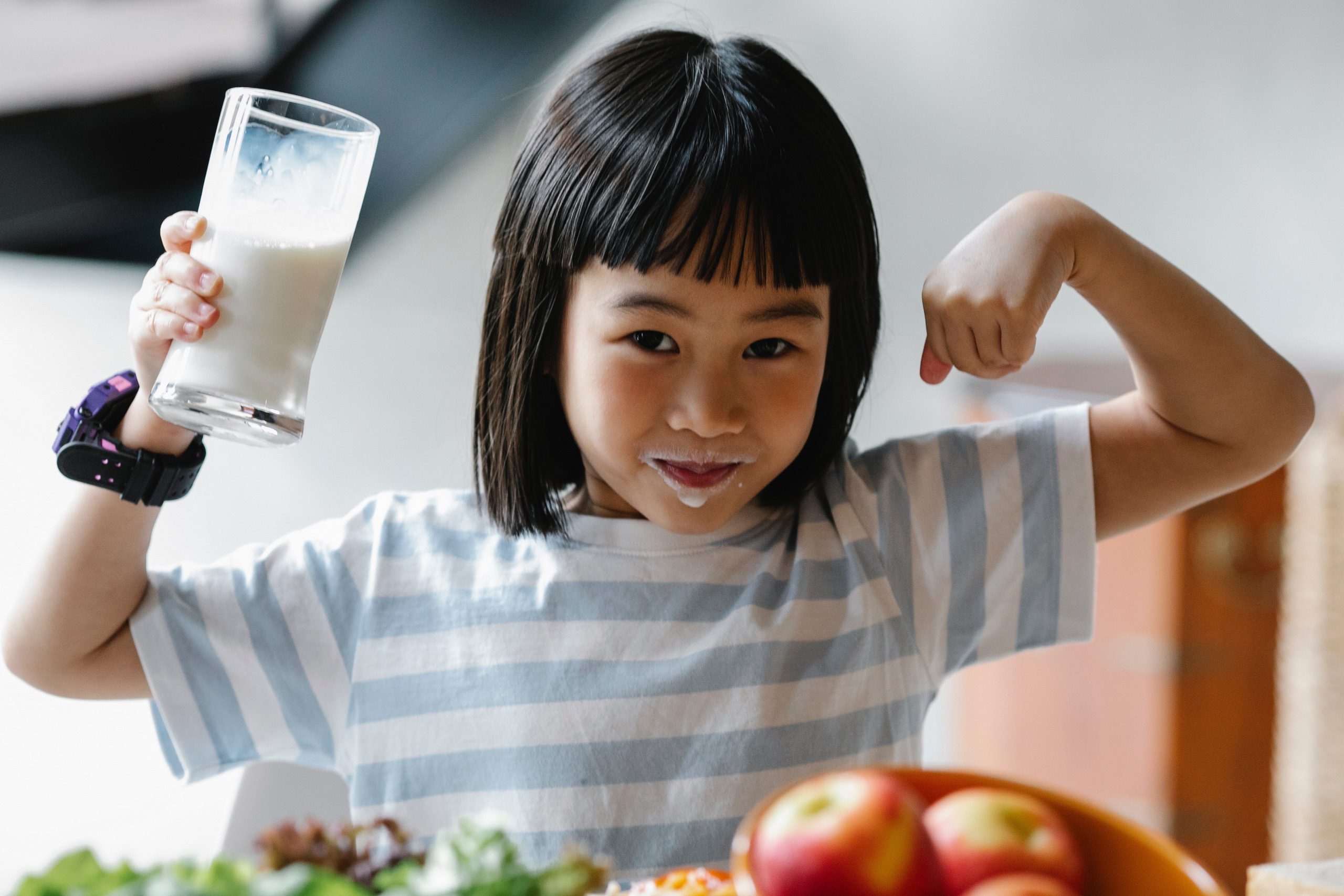 A young girl in a blue and white striped t-shirt. She is holding a glass of milk and has milk around her mouth, and has riased her arms up to show her muscles