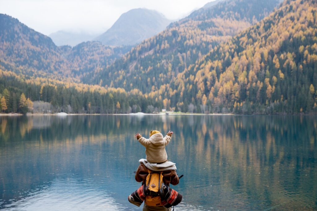 A small child on the shoulders of an adult, facing out over a large lake and some mountains covered in trees