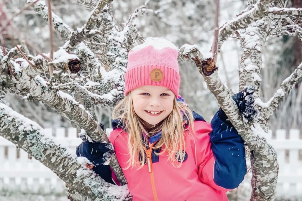 A little girl standing in the branches of a tree that is covered in snow. She is wearing Polarn O. Pyret ski gear