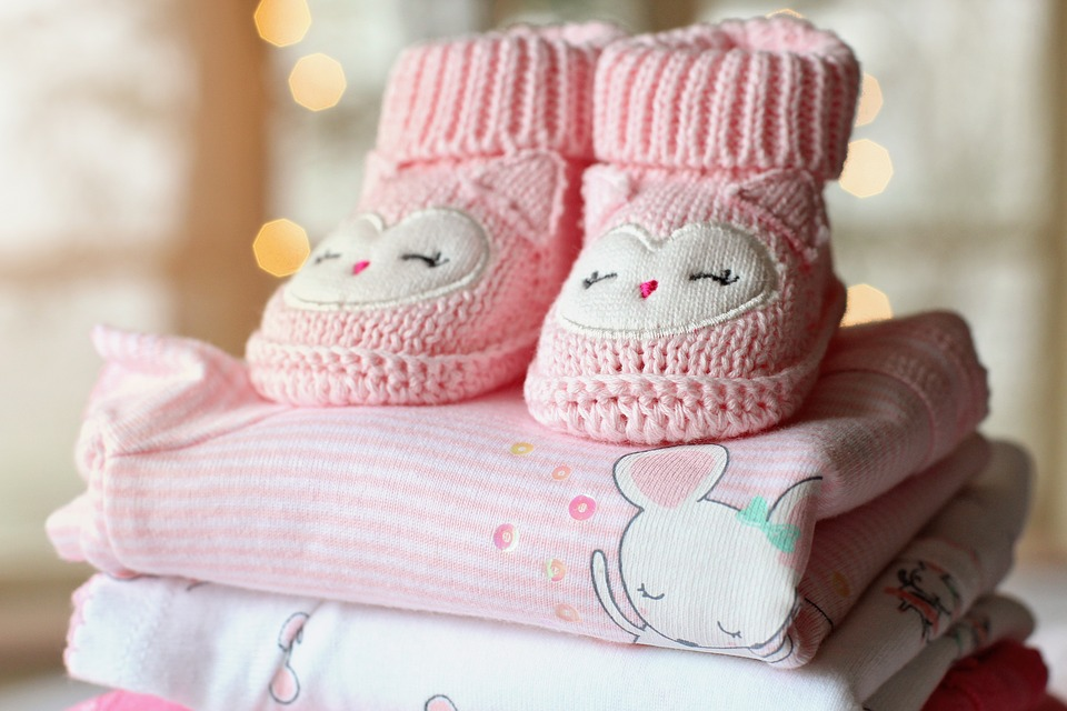 Pink baby booties sit on a pile of folded pink and white baby clothes.