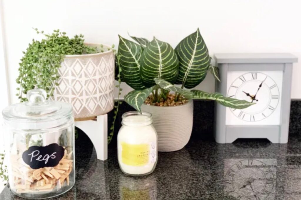 A kitchen counter with a small clock, plants, a candle and a jar on it