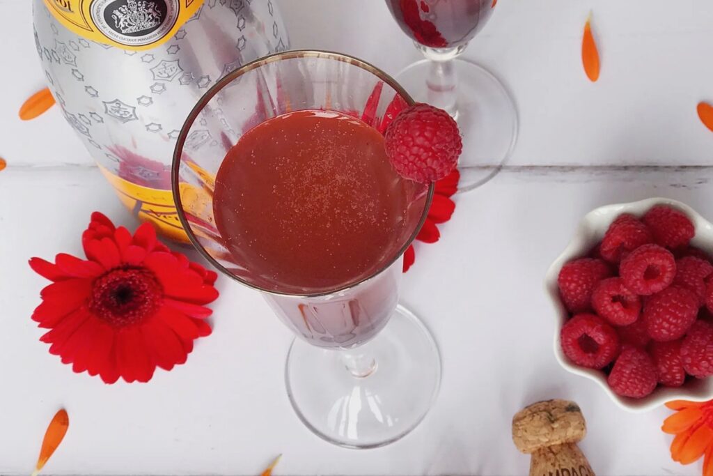 A red cocktail in a champagne glass. A bowl of raspberries is next to it alone with a bottle of champagne and some flowers